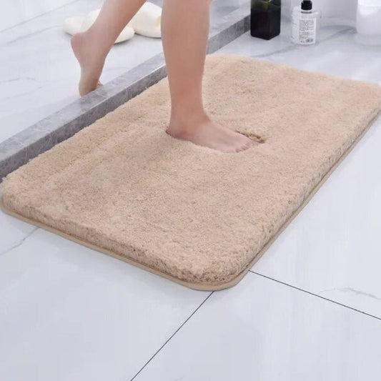 A beige plush bath mat with a deep face and a bound edge, on a white tiled bathroom floor, with a bare foot sunk into it.