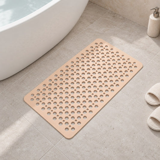 A beige non slip shower mat lying flat on a limestone bathroom floor beside a white freestanding bath, its round drainage holes visible across the whole mat