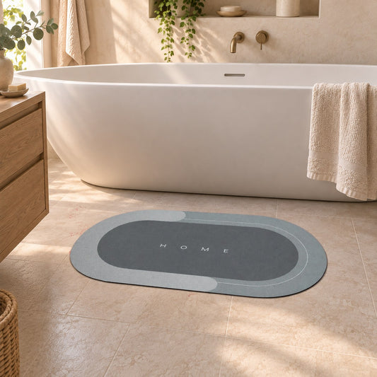 A gray oval bath mat with a pale border and the word Home printed small in the middle, lying on the tiled floor of a warm bathroom beside a freestanding bath.