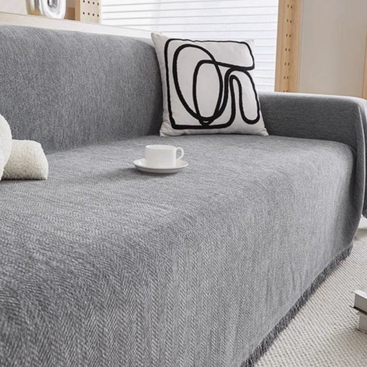 A gray chenille cover spread over a couch in a bright room, with a woven herringbone face and a tassel fringe along the front edge.