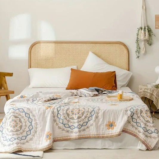 A cream mandala throw blanket spread over a made bed with a rattan headboard and an orange bolster cushion.