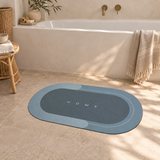 A blue oval bath mat with a pale border and the word Home printed small in the middle, on the floor of a bathroom with limewashed walls and a rattan stool.