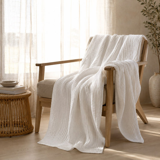 White muslin cotton blanket draped over a pale oak armchair and backlit by a window, so the open weave shows against the light