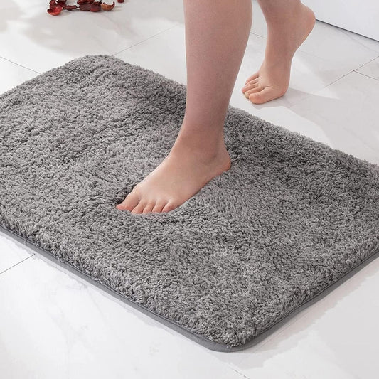A gray plush bath mat on a white marble floor, with a bare foot sunk into it.