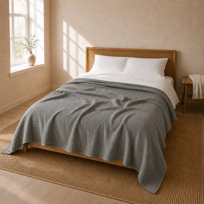 Gray muslin cotton blanket thrown across a bed with a woven rattan headboard in a bright guest room, the whole bed and the room in view