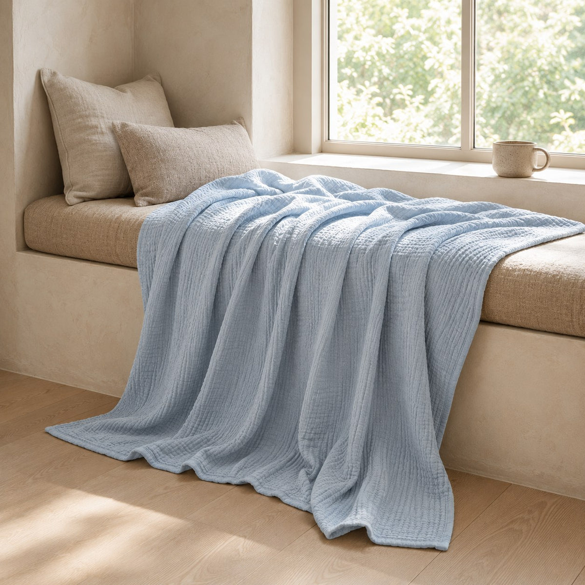 Pale blue muslin cotton blanket spilling off a window seat onto a pale oak floor in bright daylight