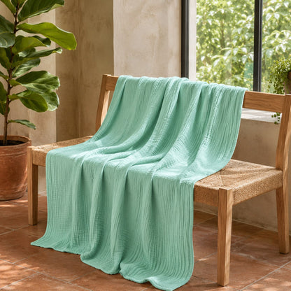Jade green muslin cotton blanket over a woven oak bench in a garden room with terracotta tiles and plants