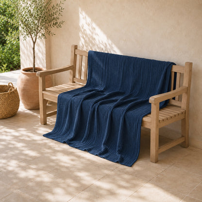 Denim blue muslin cotton blanket opened out along an oak bench on a sunlit covered porch, falling over the front edge onto the stone floor