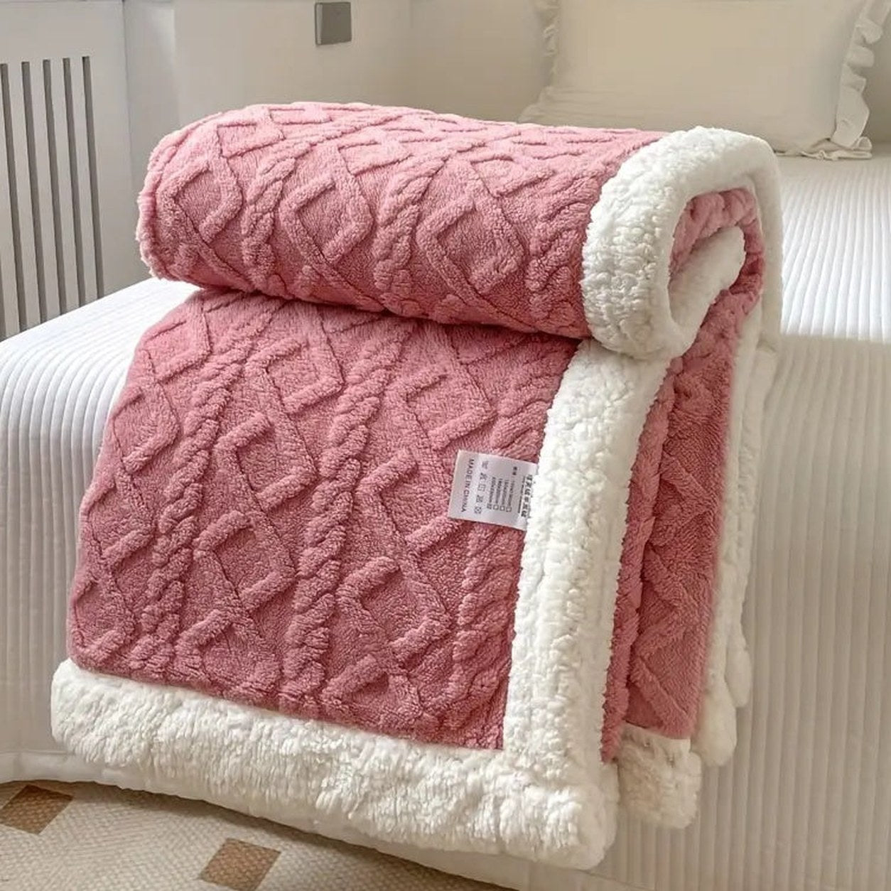 Pink sherpa throw blanket folded on a bed, the raised cable texture facing out and the white sherpa lining showing at the fold