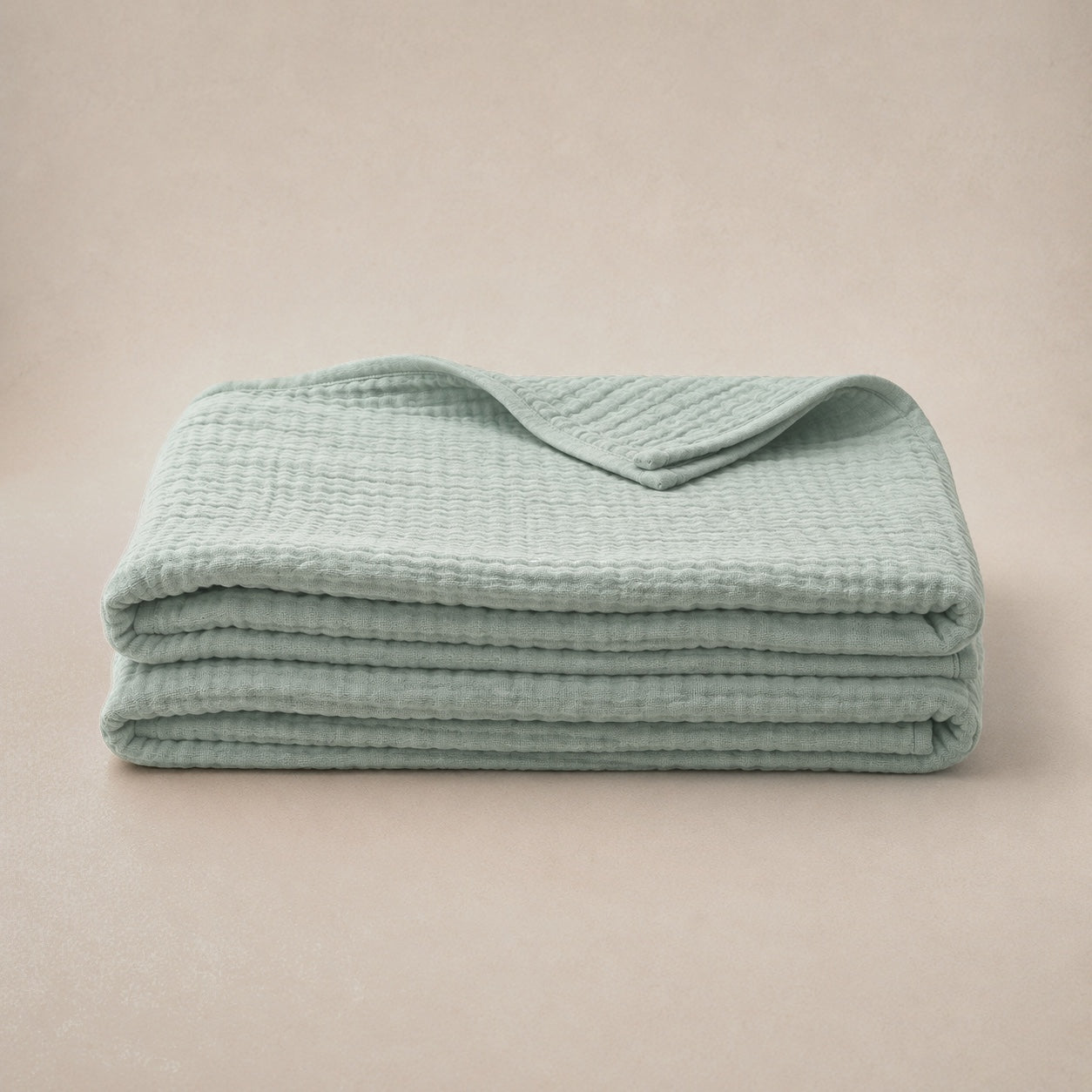 The sage muslin cotton blanket folded into a stack on a warm plaster studio backdrop, with the top fold turned back to show the layered edge
