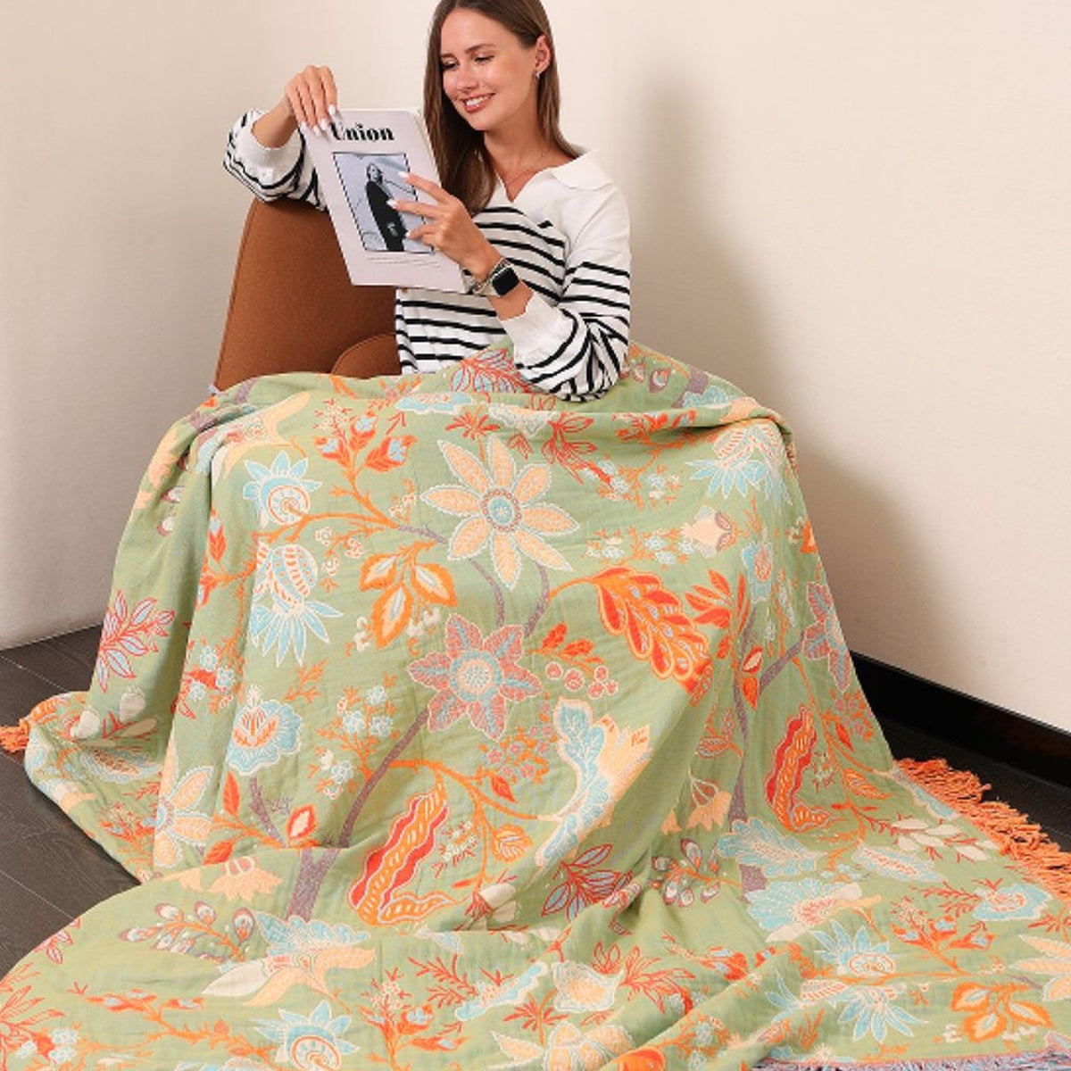 The sage green face of the floral cotton throw drawn over a woman's lap in a chair and spilling to the floor, the tassel fringe along the edge.