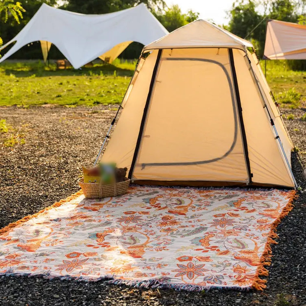 The floral cotton throw spread flat on the ground outside a tent in late afternoon sun, cream face up with the tassel fringe running along all four sides.