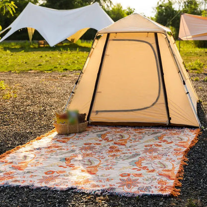 The floral cotton throw spread flat on the ground outside a tent in late afternoon sun, cream face up with the tassel fringe running along all four sides.